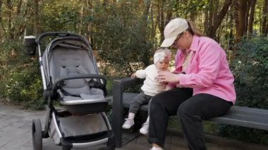 Baby girl and her young mother sitting on park bench and using smartphone