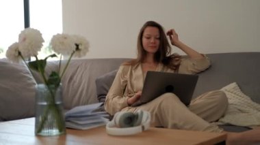 Concentrated young woman in bathrobe working on laptop while sitting on cozy sofa at home with fresh flowers in vase