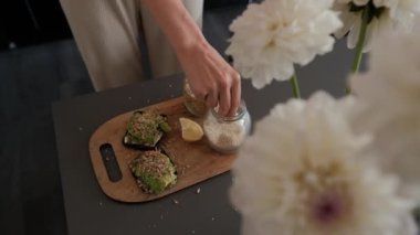 Woman sprinkles avocado toast with sesame seeds and pours lemon juice over it on table with a beautiful bouquet of flowers