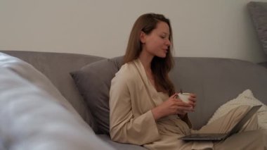 Side view of young woman in bathrobe is sitting on sofa at home drinking coffee and making video call using laptop