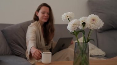 Blurred woman in a bathrobe is drinking coffee and using a laptop, focus on a fresh flowers in vase on foreground