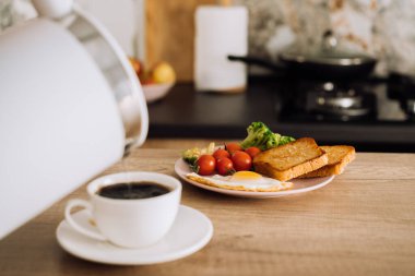 Homemade breakfast on the wooden table in the kitchen, blurred cup of coffee with kettle on foreground