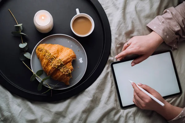 Flat lay of woman using digital tablet for drawing while lying on bed and enjoying croissant with cappuccino