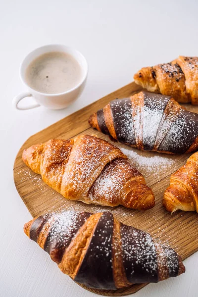 Bunch of appetizing brown and chocolate croissants with powdered sugar on wooden board with cup of coffee on white table