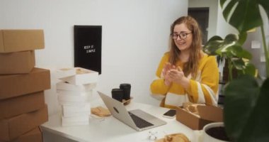 Happy woman wearing eyeglasses rejoices while working on laptop among boxes with goods, excited entrepreneur starting small business at home office