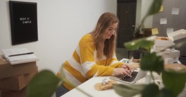 Entrepreneur female starting small business in her apartment at home, young woman working on laptop in kitchen among boxes with goods
