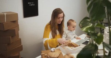 Entrepreneur woman with baby child on lap working at office among boxes of goods, young mother starting small business at home