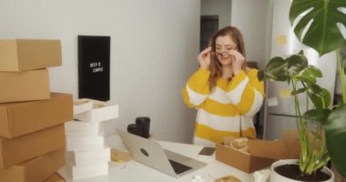 Genç bir kadın kutuların arasında dizüstü bilgisayarla çalışırken gözlük takıyor. Girişimci kadın evde küçük bir iş kuruyor.