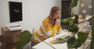 Young woman working on laptop in kitchen among boxes with goods, entrepreneur female starting small business at home