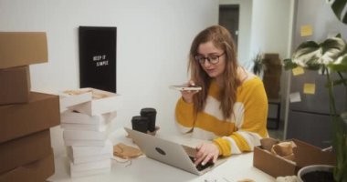 Woman in glasses using smartphone for voice mail while working on laptop among boxes with goods, entrepreneur starting small business at home