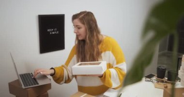 Young woman working on laptop in kitchen among boxes with goods, entrepreneur female starting small business in her apartment at home