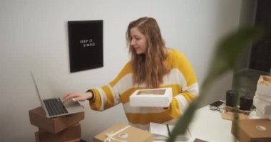 Entrepreneur female starting small business in her apartment at home, young woman working on laptop in kitchen among boxes with goods