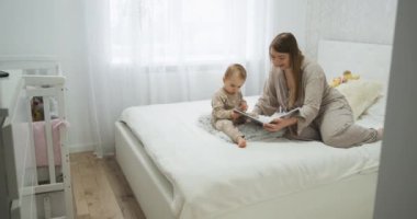 Baby child with her young mother reading a book on the bed in bedroom