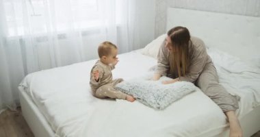 Mother in pajamas playing with baby daughter on bed, young mom with child in bedroom