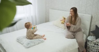Mother and her baby daughter in pajamas clapping hands on bed, young mom with child in bedroom with green plant in the foreground