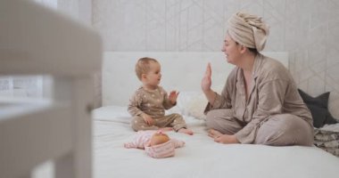 Baby child gives a high five to mother and kisses her on bed, young mom with toddler daughter in pajamas at home