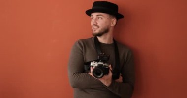 Portrait of young man with hat and camera, cheerful male photographer in studio with terracotta background