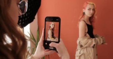 Female photographer takes a picture of a model on smartphone in the studio with terracotta background