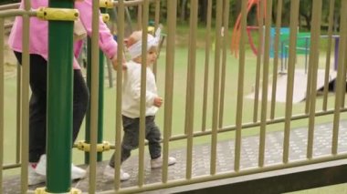 Young mother helping her baby girl with a bow on her head making first steps, little child with mom on the playground. High quality 4k footage