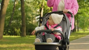 A cheerful little girl in a baby carriage, a young mother drinks coffee while walking with her child in an outdoor park. High quality 4k footage