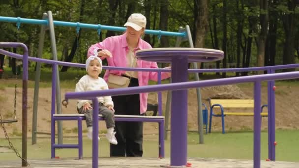 若い母親は屋外公園の遊び場でカルーセルに娘を乗せています 高品質4K映像 — ストック動画