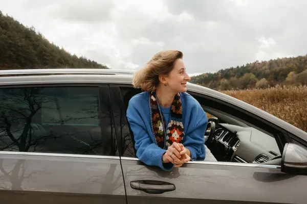 A woman leans out of car window during road trip in autumn, the wind blows her hair