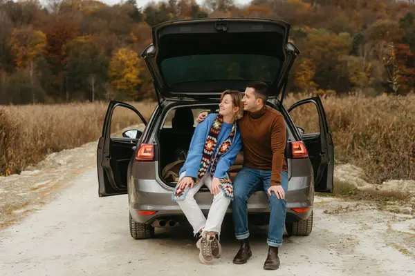 Happy young woman and man sitting in open trunk of their car while traveling in autumn and looking away, couple road trip concept