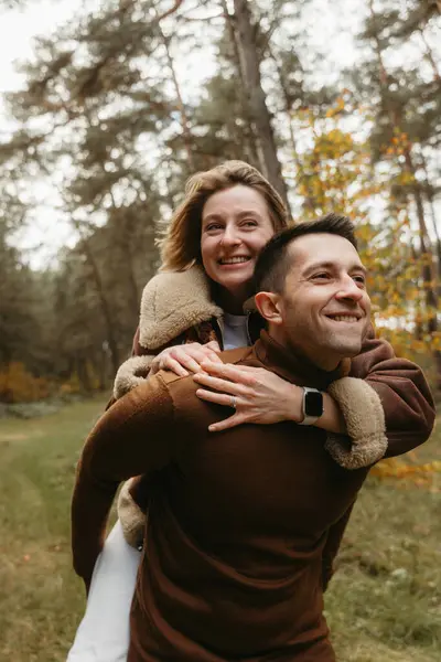 在外面的秋天公园里玩得很开心的一对成年夫妇 快乐的男人背着女人在树林里 图库照片