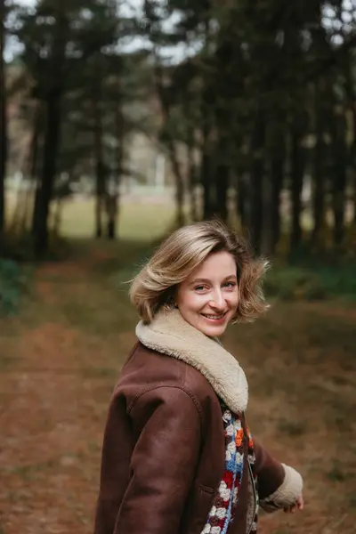 穿着棕色夹克的快乐快乐的成年高加索女人正走在秋天的森林里 图库图片