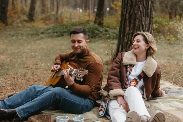 一个男人正在秋天的树林里为一个快乐的女人弹奏吉他 而她却坐在毛毯上 免版税图库图片