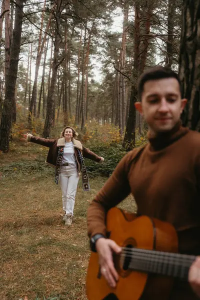 男人在森林里弹吉他 女人在秋天的公园里跟在他后面跳舞 图库照片
