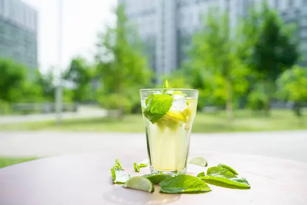 glass with fresh spring or summer iced drink mocktail with lemon and mint at table in caffe terrace in day light in front of trees and buildings. High quality photo