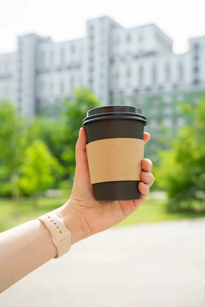 womans hand holding black hot cup of coffee tea in front of buildings trees and sky. High quality photo