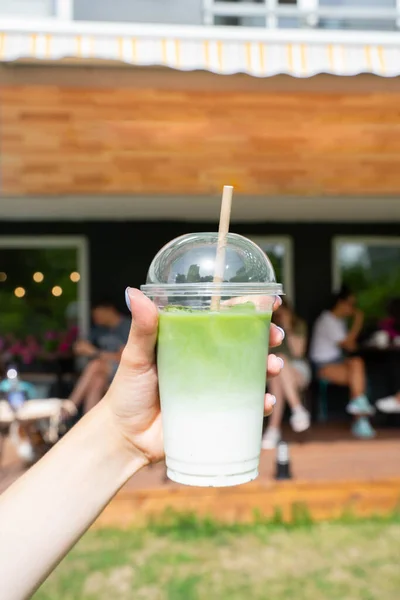Womans hand holding cold brewed iced matcha latte with ice in plastic cup with straw upfront cafe terrace. High quality photo