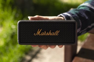 Güneşli bir günde siyah Marshall taşınabilir bluetooth hoparlörü tutan birinin yakın çekimi. İkonik markalaşma ve doğal bulanık arkaplan ile modern kablosuz ses aygıtı