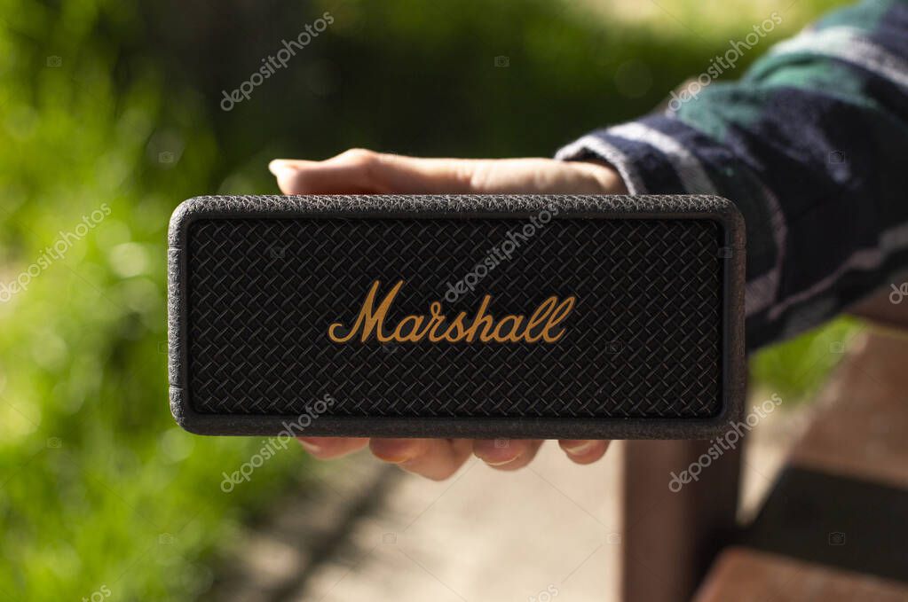 Close-up of a person holding a black Marshall portable Bluetooth speaker outdoors on a sunny day. Modern wireless audio device with iconic branding and a natural blurred background