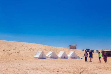 Bir grup insan, Mısır 'ın altın çölünde, canlı mavi bir gökyüzünün altında kurulmuş beyaz çadır sırasının yanında yürüyorlar. Macera, turizm, eko seyahat ya da kamp hayatı için mükemmel bir konsept.