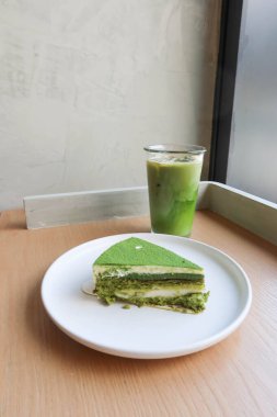 Japon kibrit peynirli peynirli keki beyaz bir tabakta mochi, buzlu yeşil çaylı latte ile ahşap bir masada, doğal güneş ışığıyla bir kafede çekilmiş.