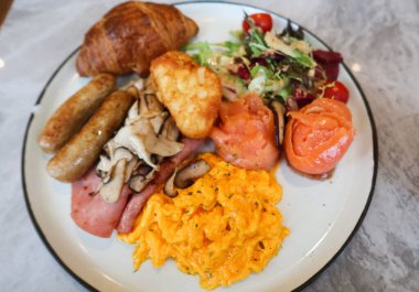 İngiliz kahvaltısını çırpılmış yumurta, tütsülenmiş somon rulosu, salata sebzesi, domuz sosisi, jambon, kızarmış mantar, beyaz tabakta haşlanmış kahverengi ve kruvasan ile kapatın.