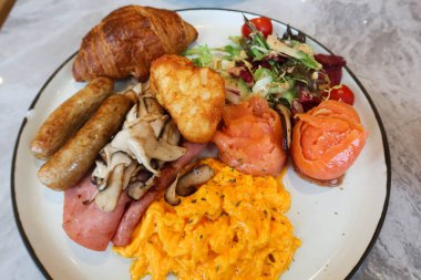 İngiliz kahvaltısını çırpılmış yumurta, tütsülenmiş somon rulosu, salata sebzesi, domuz sosisi, jambon, kızarmış mantar, beyaz tabakta haşlanmış kahverengi ve kruvasan ile kapatın.