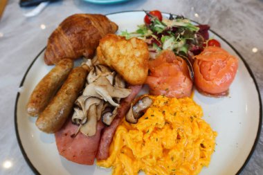İngiliz kahvaltısını çırpılmış yumurta, tütsülenmiş somon rulosu, salata sebzesi, domuz sosisi, jambon, kızarmış mantar, beyaz tabakta haşlanmış kahverengi ve kruvasan ile kapatın.