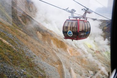 Owakudani, Japonya- 29 Ekim 2024: dağ boyunca uzanan duman ve teleferik halatıyla volkan dağı farklı renklerde kayalar, kahverengi ve turuncu yol manzaralı.  