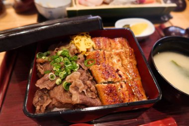 Japon yemek çantasını (bento) yarım ızgara yılan balığı pilavı ve yarım wagyu bifteği ile kapatın. Miso çorbası yanında, başlangıç ve kırmızı kaşık.