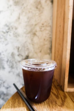 Şeffaf plastik fincana ve kafede çiğ çimento duvarı ve ahşap masası olan siyah buzlu Americano sade kahvesine yakın dur.  