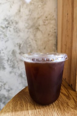 Şeffaf plastik fincana ve kafede çiğ çimento duvarı ve ahşap masası olan siyah buzlu Americano sade kahvesine yakın dur.  