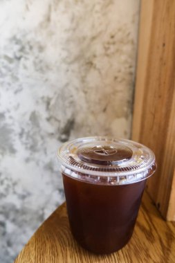 Şeffaf plastik fincana ve kafede çiğ çimento duvarı ve ahşap masası olan siyah buzlu Americano sade kahvesine yakın dur.  