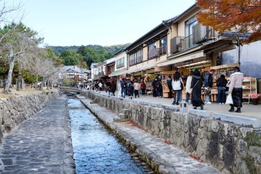 Miyajima, Hiroşima, Japonya - 10 Aralık 2024: Bir çok insanla birlikte turistik bölgede, nehir kenarında hediyelik eşya satan mağazalar 
