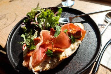 Parma jambonlu ve ekşi ekmekli tostun yanında siyah tabakta salata olsun.