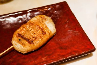 Kırmızı tabağa tavuk derisi sarılmış ızgara mürekkep balığı şişi.