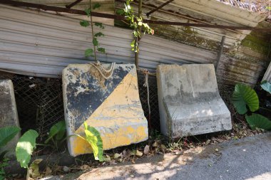 İki yıpranmış beton trafik bariyeri bir çite yaslanmış, kısmen büyümüş bitki örtüsüyle gizlenmiş. Bir bariyer soluk sarı ve siyah üçgen bir dizayn içerirken, her ikisi de işaretleri gösterir 